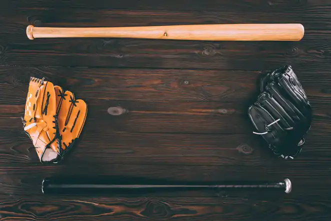 A wooden baseball bat, a black glove, and an orange glove are arranged on a rustic wooden surface, representing the gear often seen around MLB games, prompting curiosity like 'How much MLB ball boys girls make.